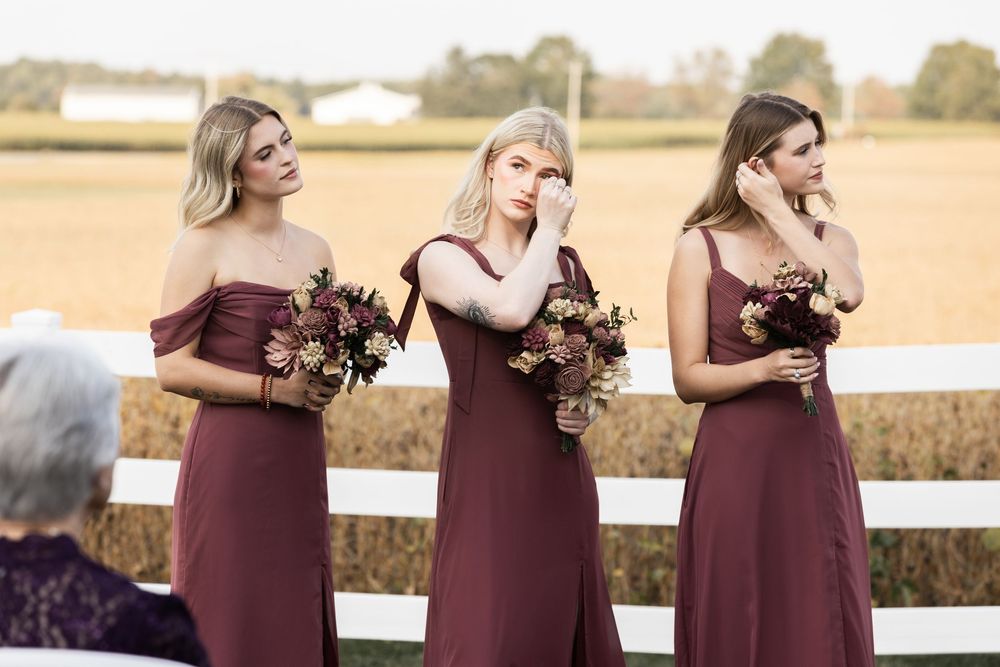 Bridesmaids in burgundy dresses stand in a line outdoors against a golden sunset field.