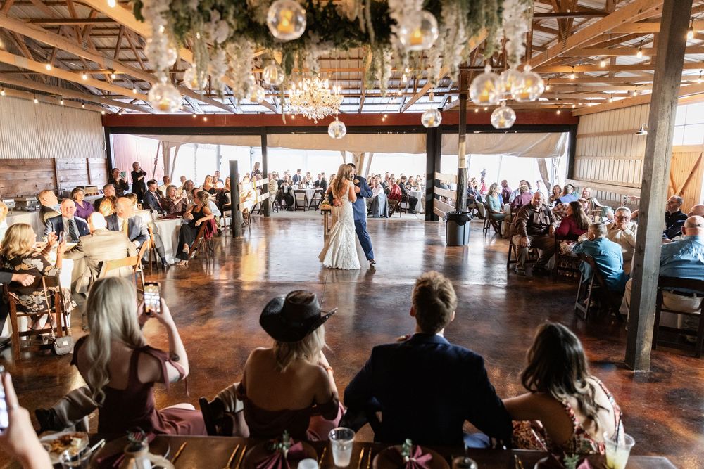 Large reception hall filled with guests watching a celebration under hanging string lights and disco balls.