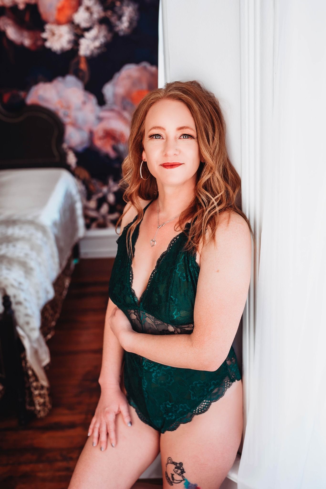 A boudoir photoshoot featuring emerald green lingerie against a white wall with pink floral decorations.
