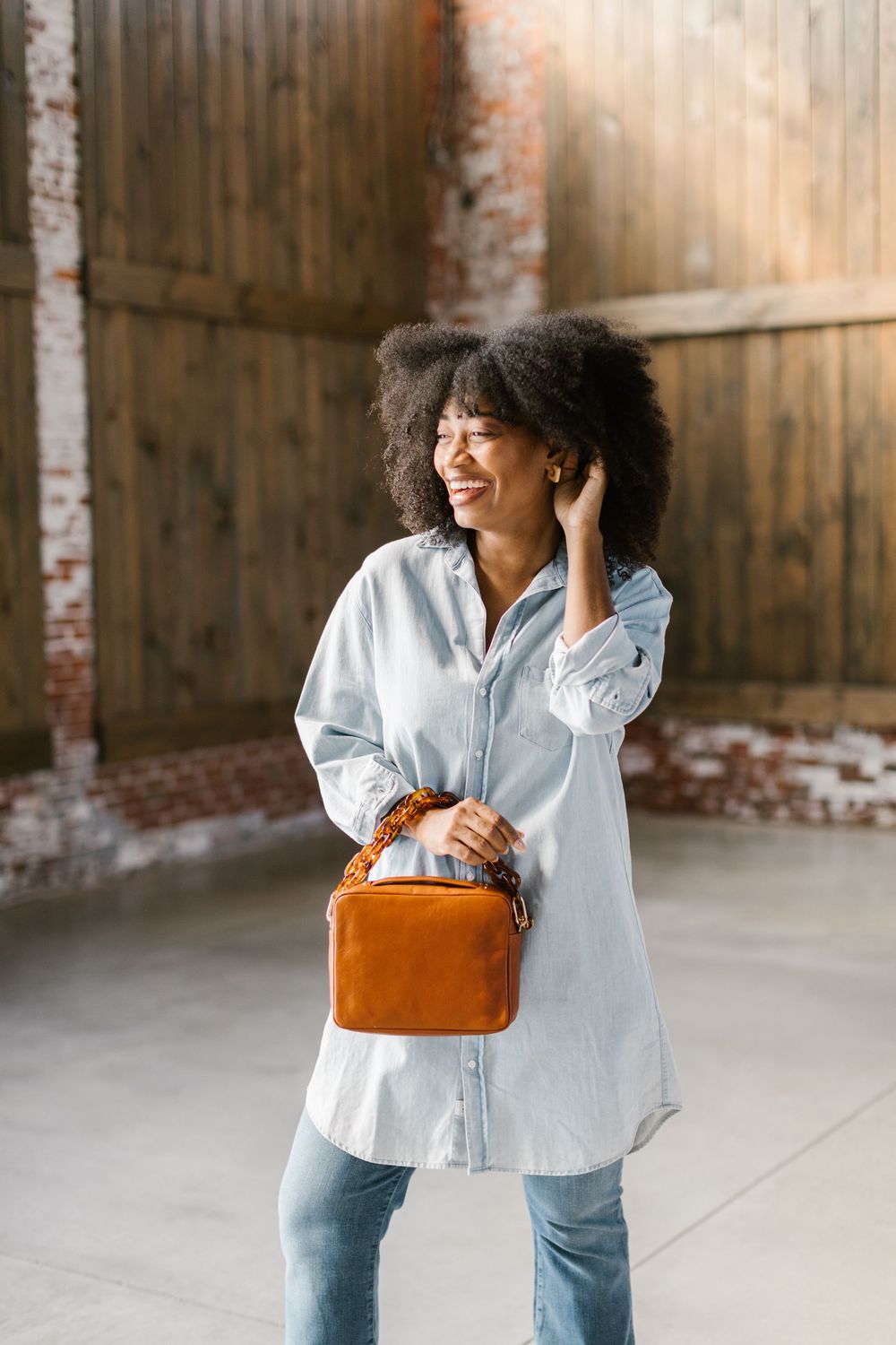 Person in light denim outfit poses playfully with orange handbag against industrial backdrop.