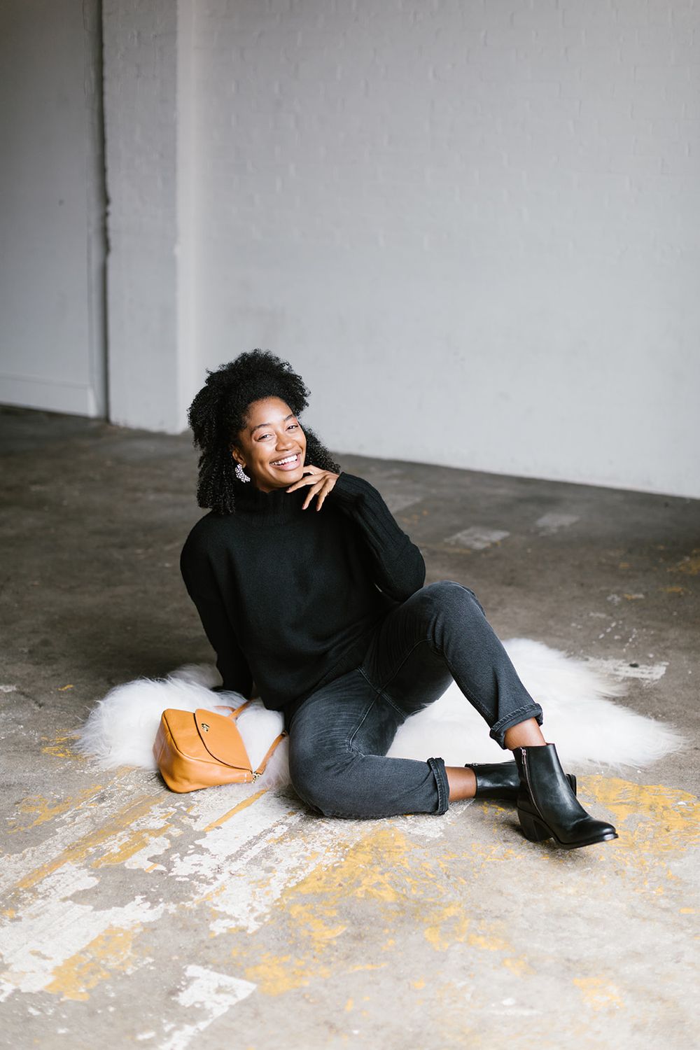 Person in black outfit reclines on floor with orange accessory in minimalist setting.