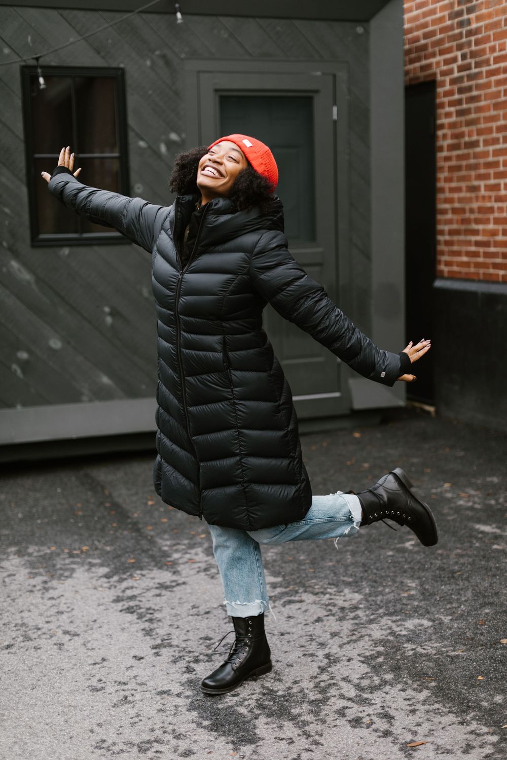 Person in black puffer jacket and jeans jumping playfully in urban setting.