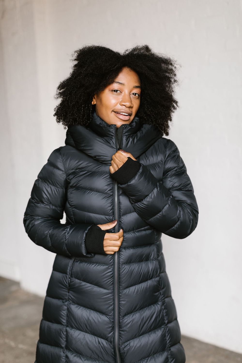 Person wearing puffy black winter coat poses against white wall in fashion photoshoot.