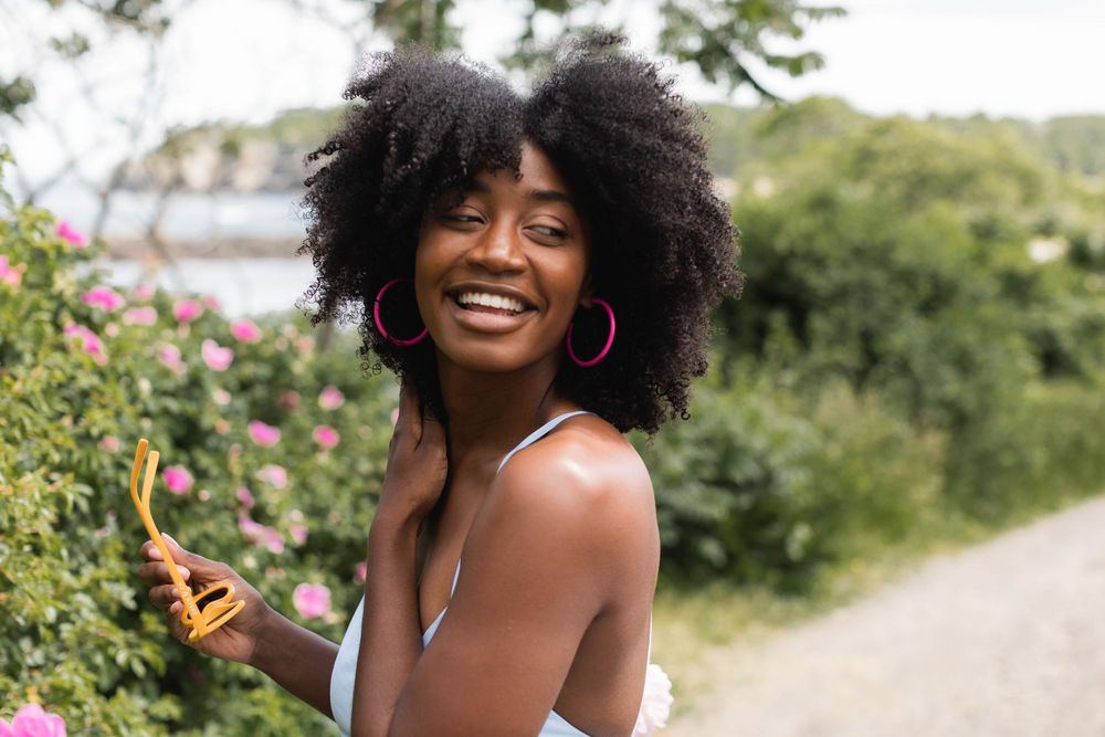 Person with natural hair poses joyfully outdoors among pink flowers and greenery.