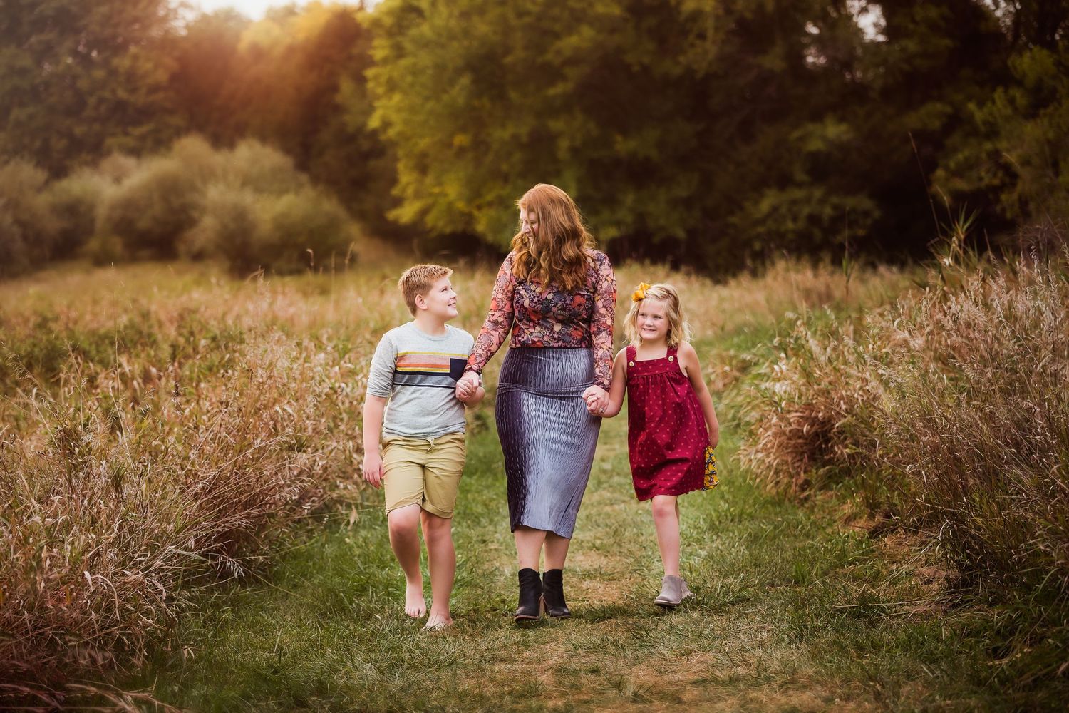 Three people walk down a scenic nature path at sunset through tall grass and wildflowers.