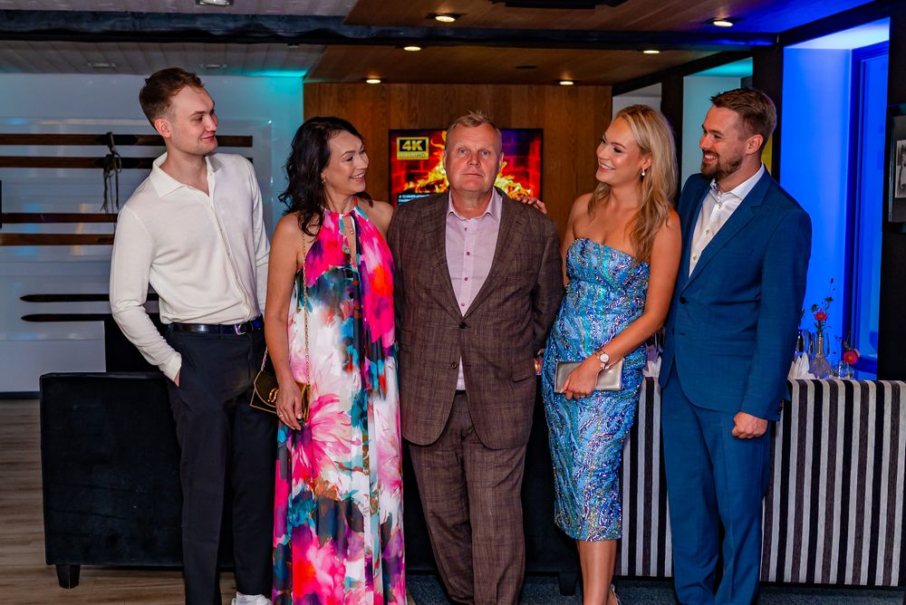 A group of people in formal attire interacting at an indoor event with blue lighting and white walls.