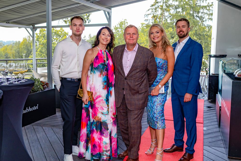 A group of five people in formal attire pose together on a covered outdoor deck during a social event.