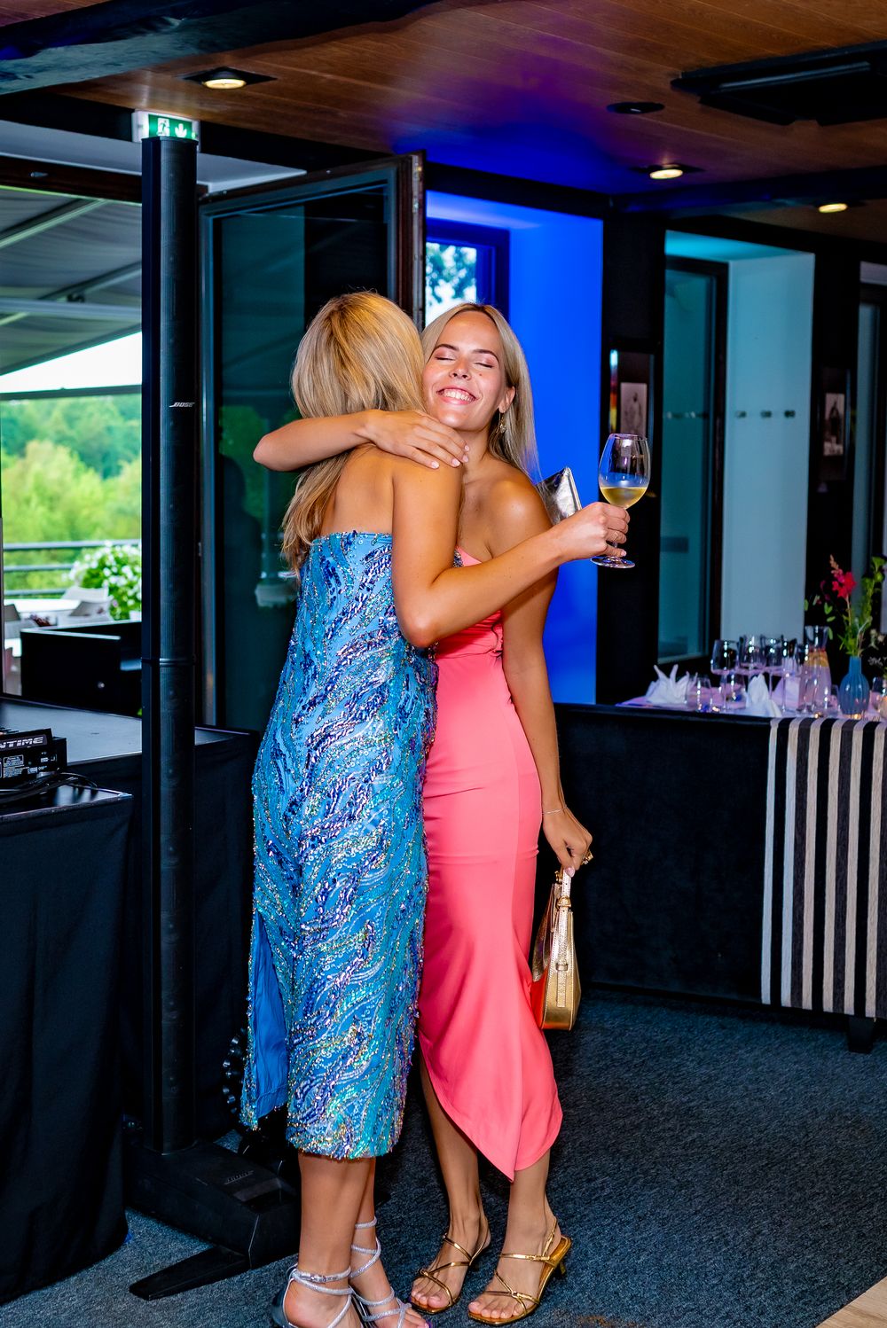 Two friends embrace while holding drinks at an indoor venue, one wearing a blue patterned dress and another in coral pink.