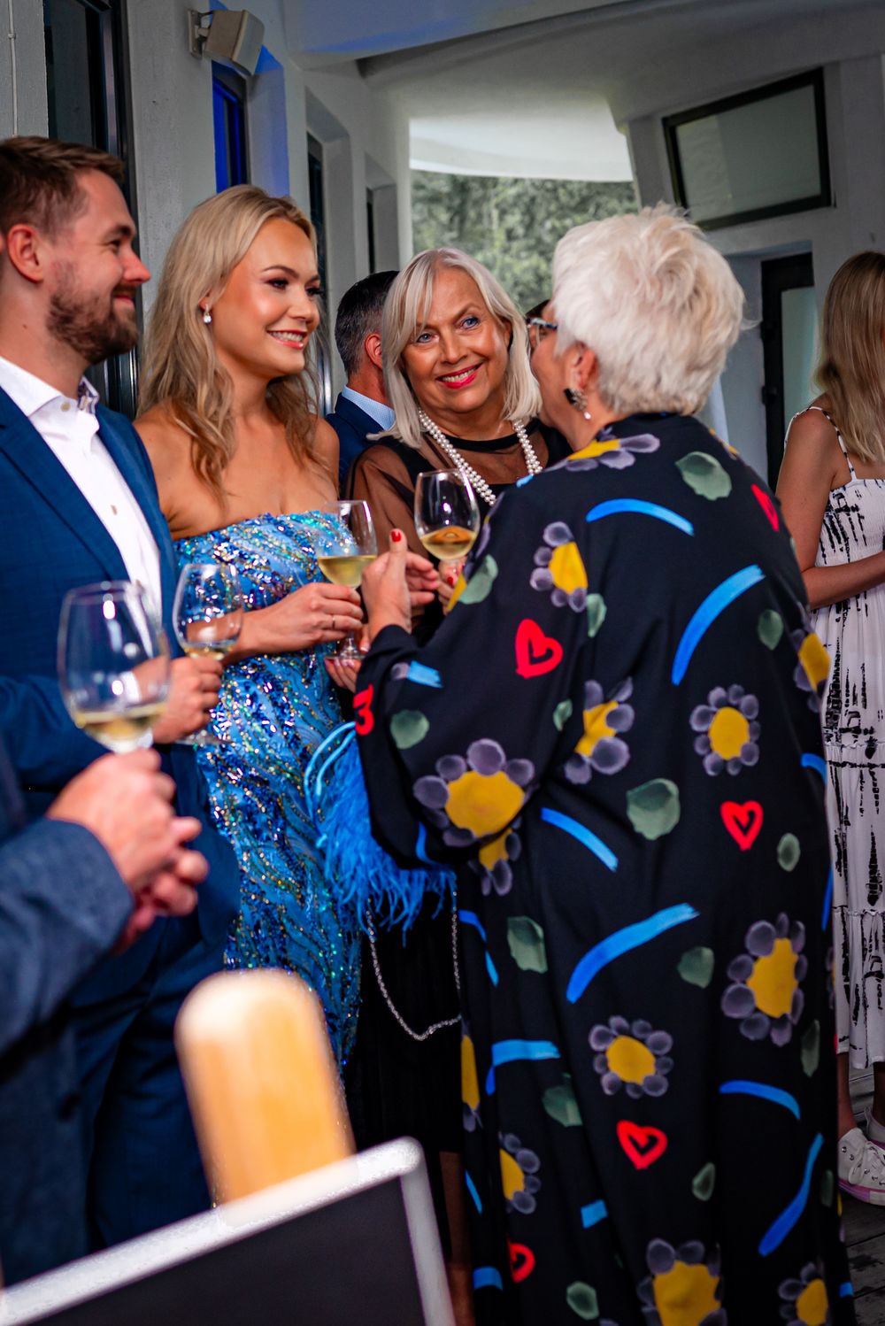 A group of people socializing at a party with wine glasses, including someone wearing a colorful patterned black shirt.