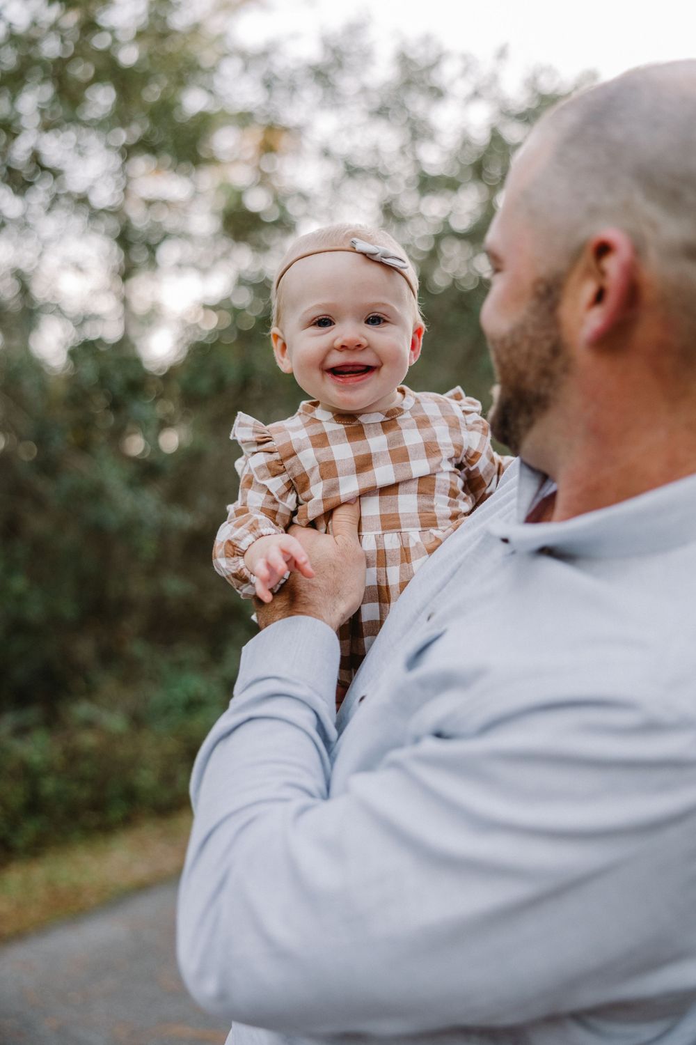 A baby wearing a brown gingham dress smiles while being held up outdoors on a sunny day.