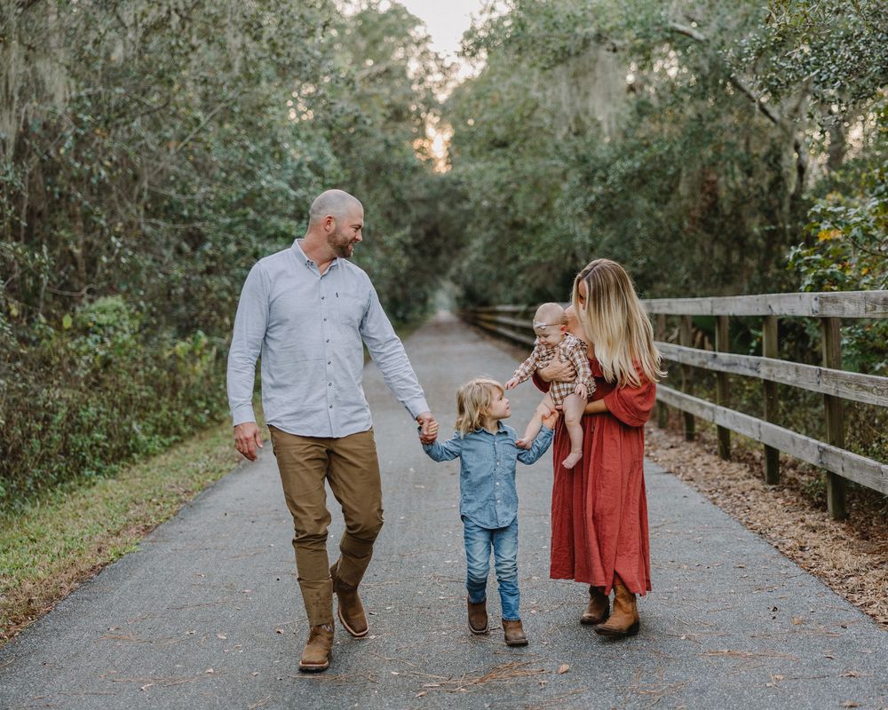 A family walks together holding hands on a tree-lined path with a wooden bridge railing on a peaceful afternoon.