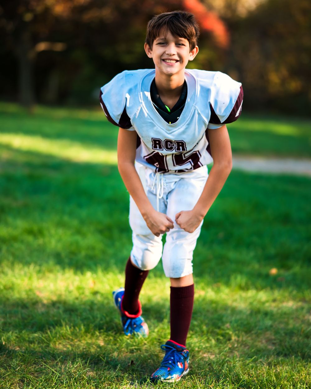 Young football player in white uniform and blue cleats crouches on sunny grass field.