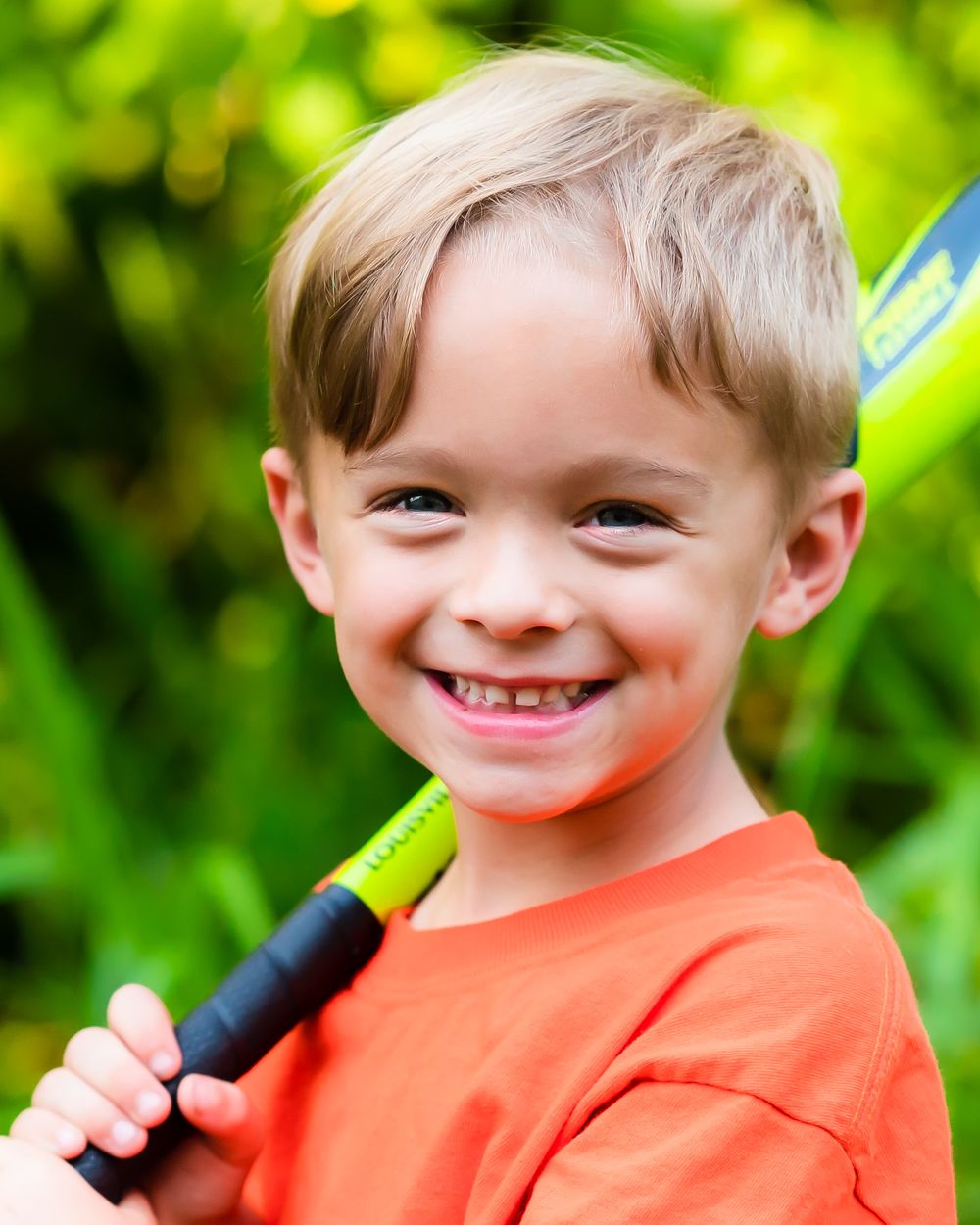 Child wearing bright orange shirt smiling against green foliage background in outdoor portrait.