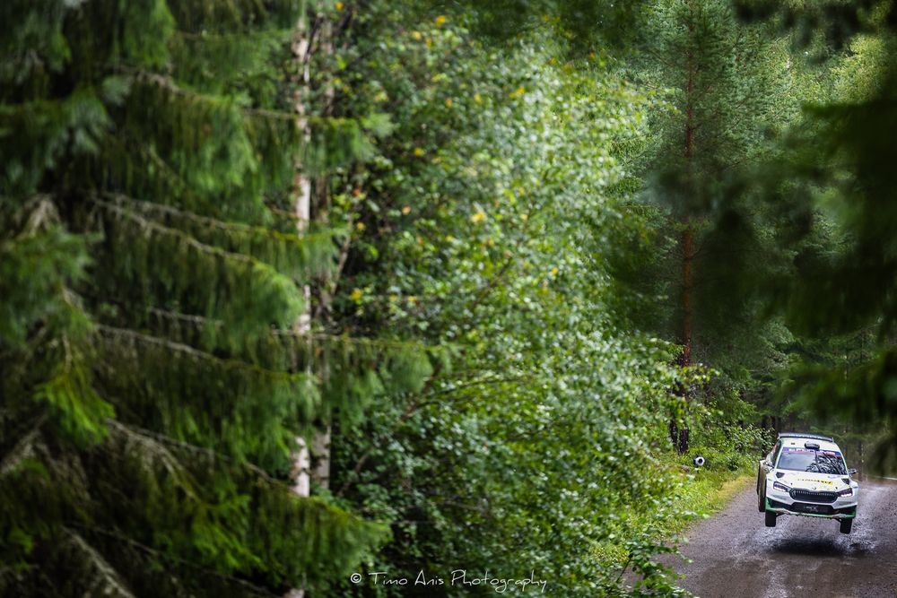 A car drives along a scenic forest road surrounded by lush green trees and vegetation.