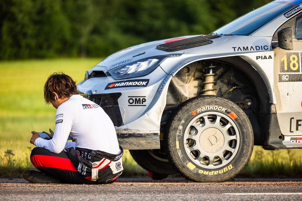 Rally car driver inspects tire damage during competitive motorsport race on gravel track.