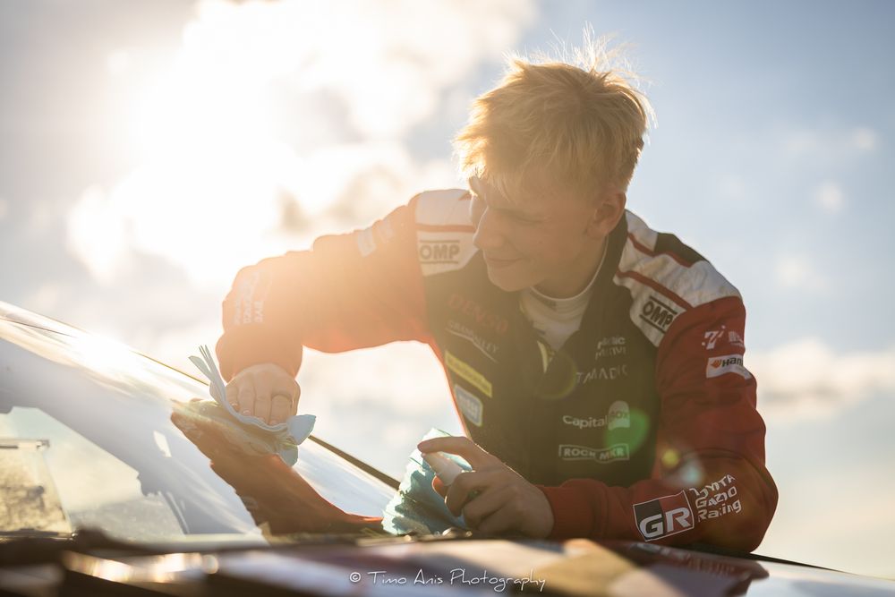 Racing driver in a black suit checks the exterior of a car during a bright sunset with lens flare.