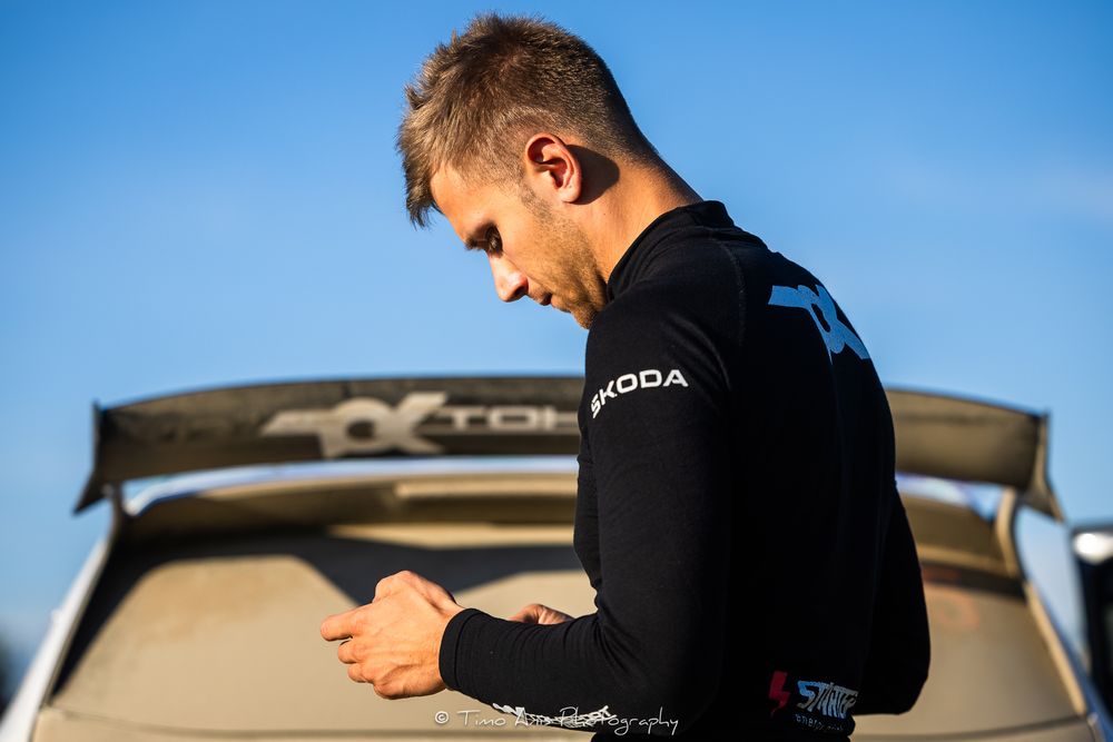 Professional surfer in a black wetsuit preparing equipment on top of a car against blue sky background.