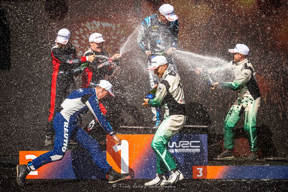 Rally drivers celebrate on podium with champagne spray during WRC victory celebration ceremony.