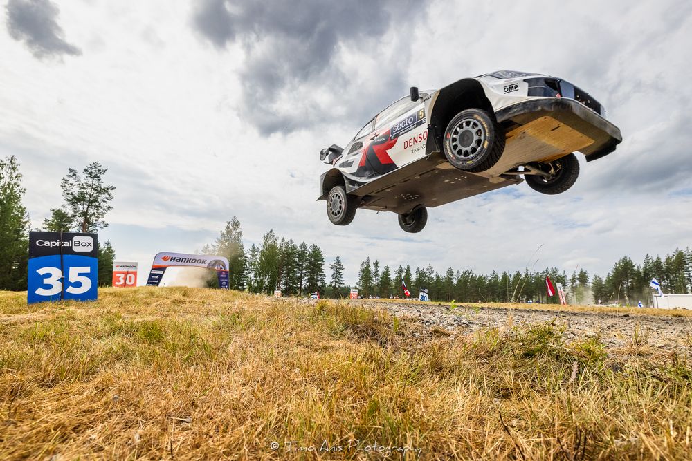 Rally car catches air while jumping over dirt hill during intense off-road racing competition.
