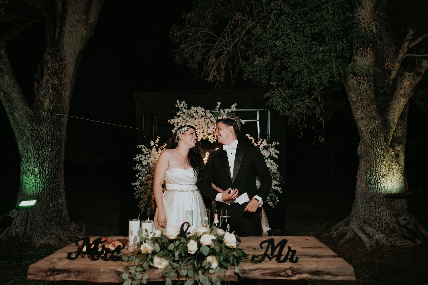 A couple walks hand in hand through a beautifully lit garden path at night with trees illuminated by soft lighting.