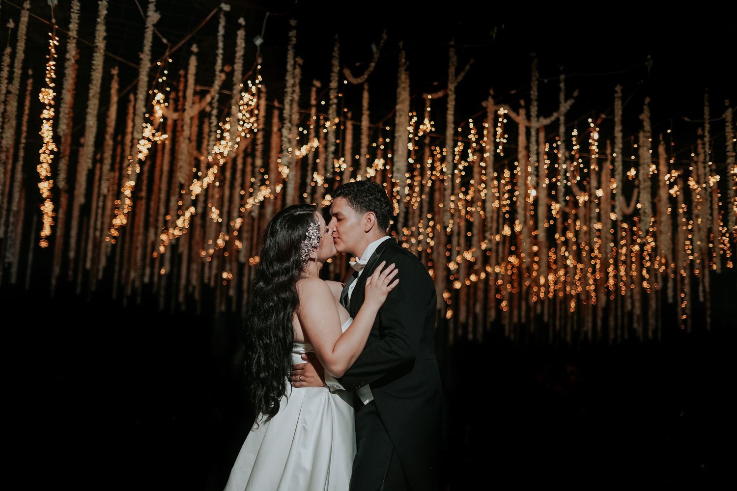 A couple shares a romantic kiss during their wedding ceremony under a glowing canopy of string lights.