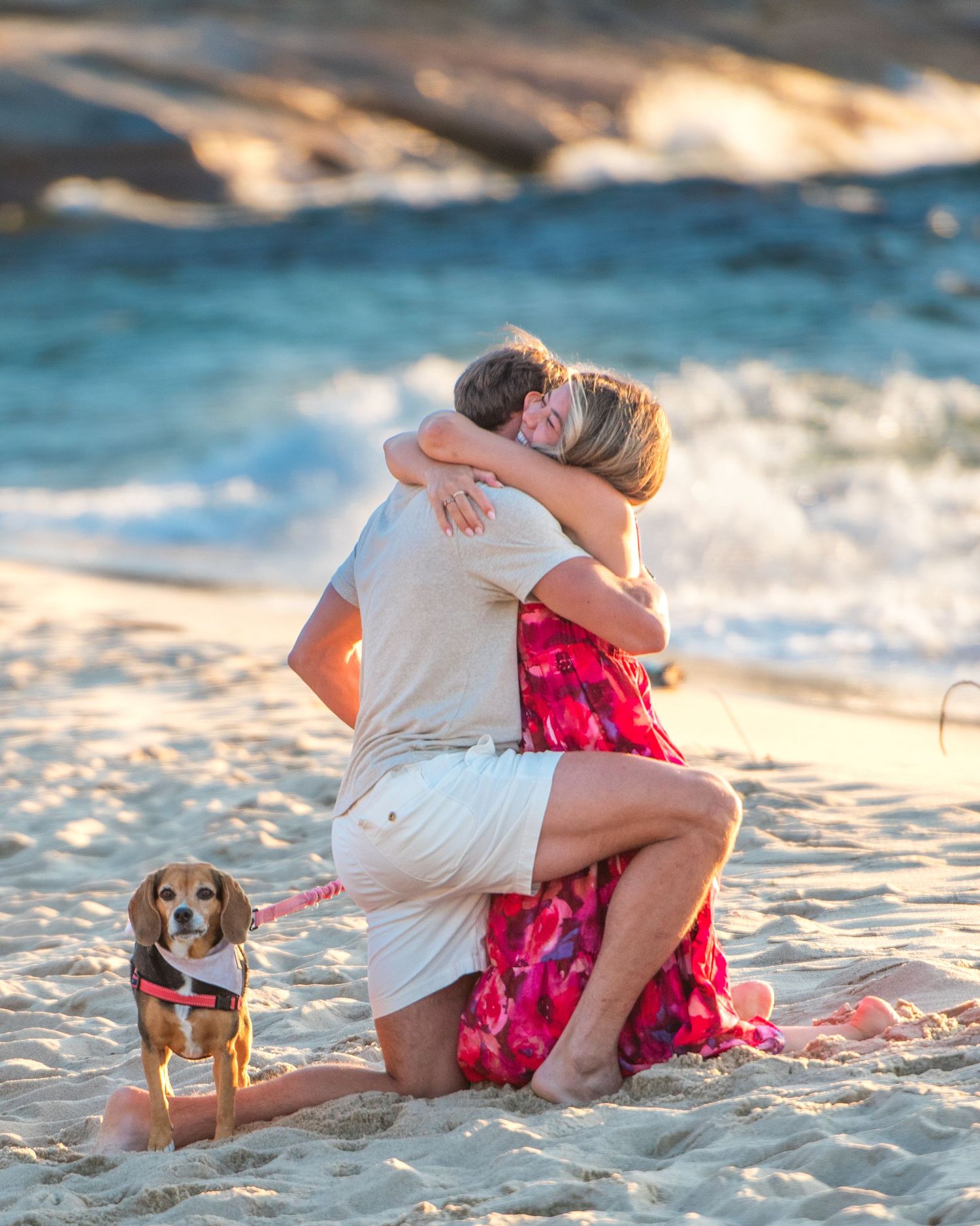 A couple embraces on a beach at sunset wearing red and black clothing while standing near the water's edge.