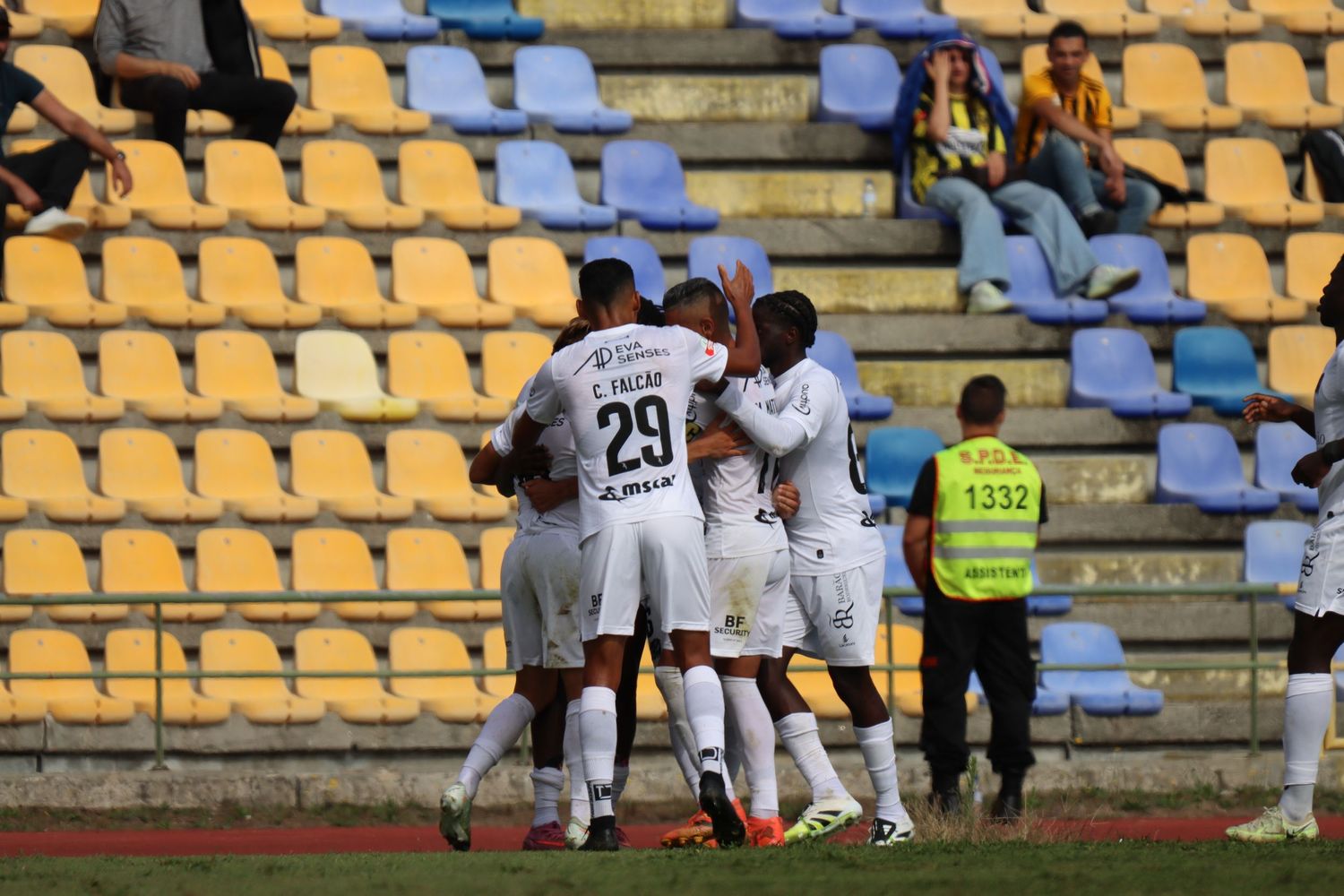 Soccer players in white uniforms celebrate together on a field with yellow stadium seats in the background.