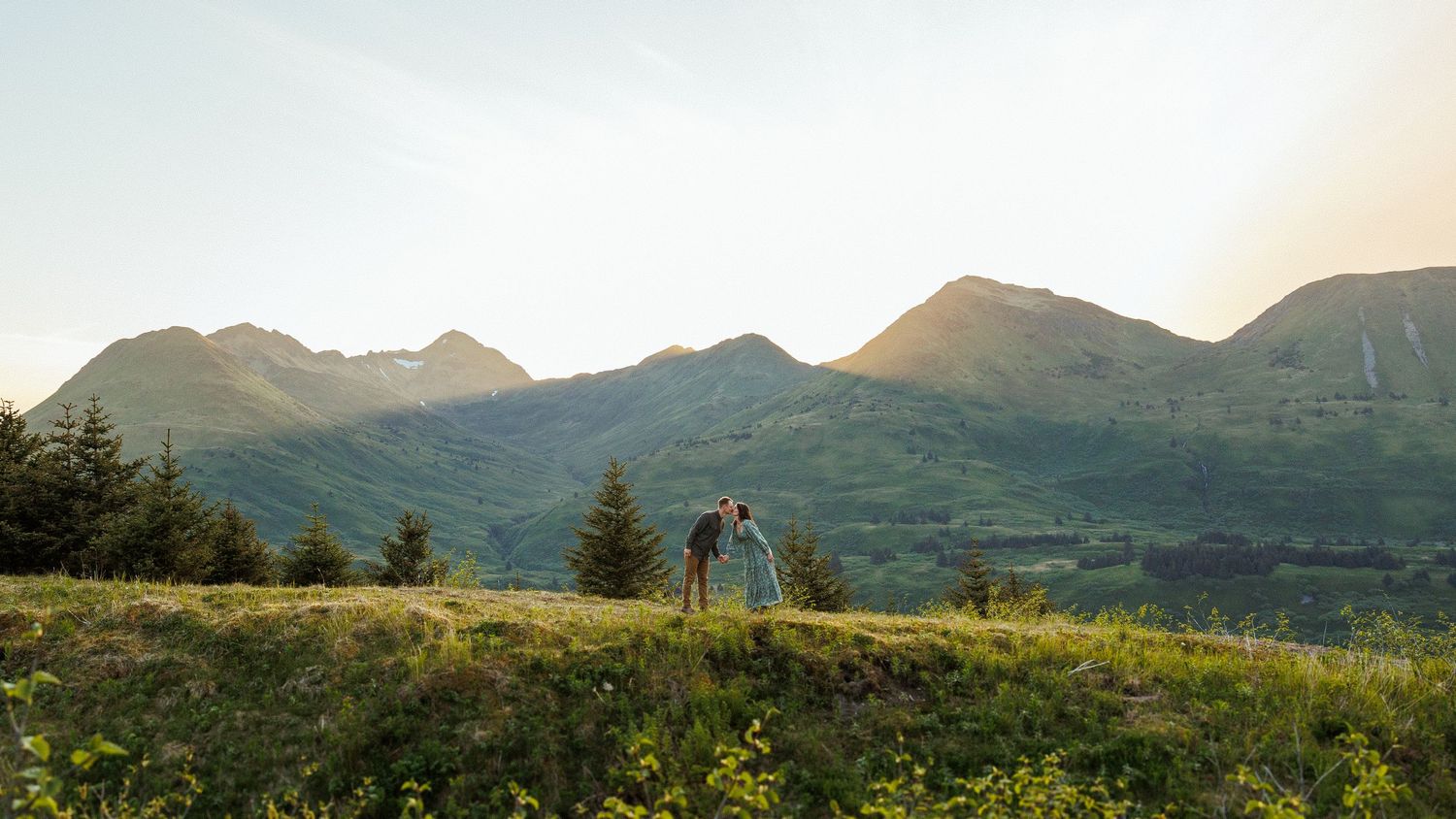 A couple stands on a mountain summit during golden hour with scenic views of lakes and mountains in the distance.