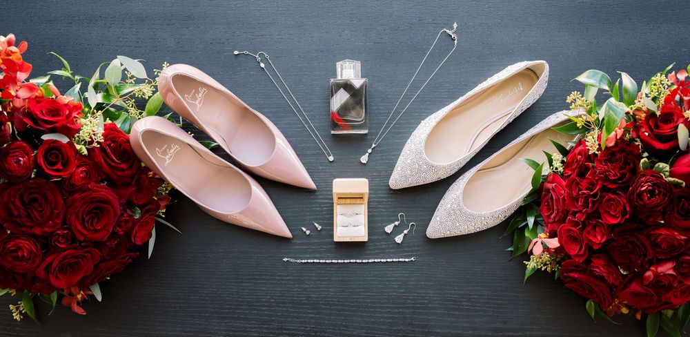 Wedding accessories including pink heels, perfume bottle, and red rose bouquet arranged on dark surface.