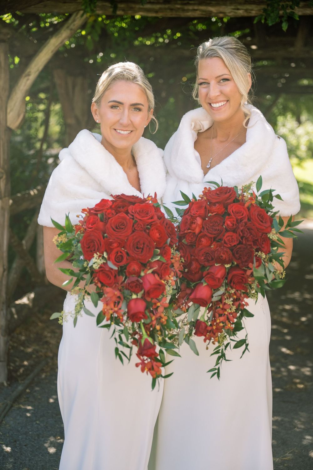 Wedding party in white dresses holding vibrant red rose bouquets outdoors.