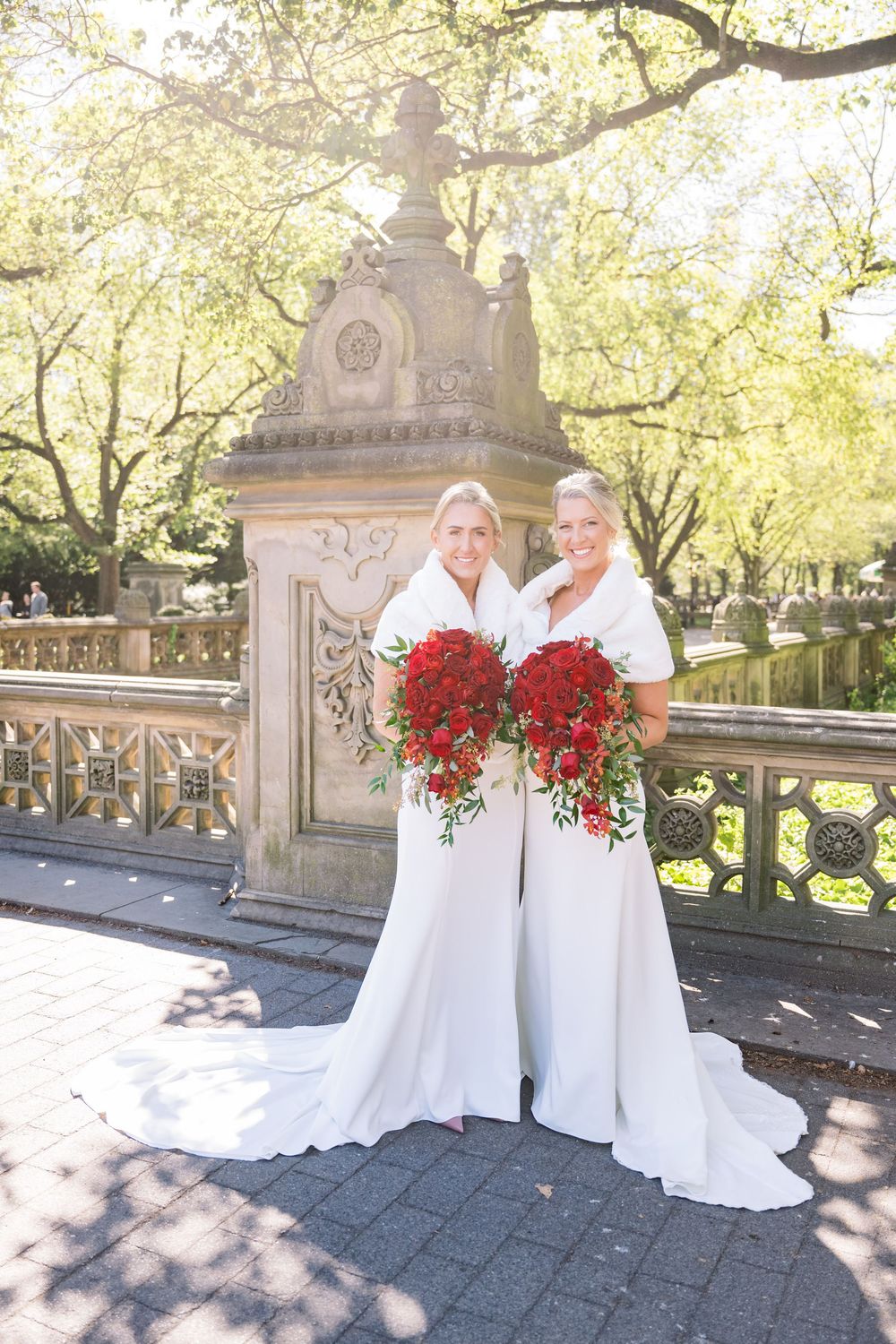 Two brides in elegant white wedding dresses hold vibrant red bouquets on ornate stone steps in a park setting.