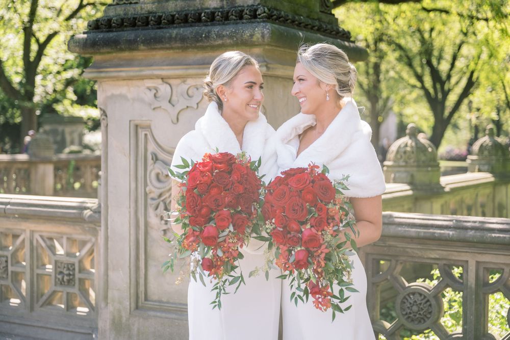 Two brides share a joyful moment while holding red floral bouquets against an ornamental stone backdrop.