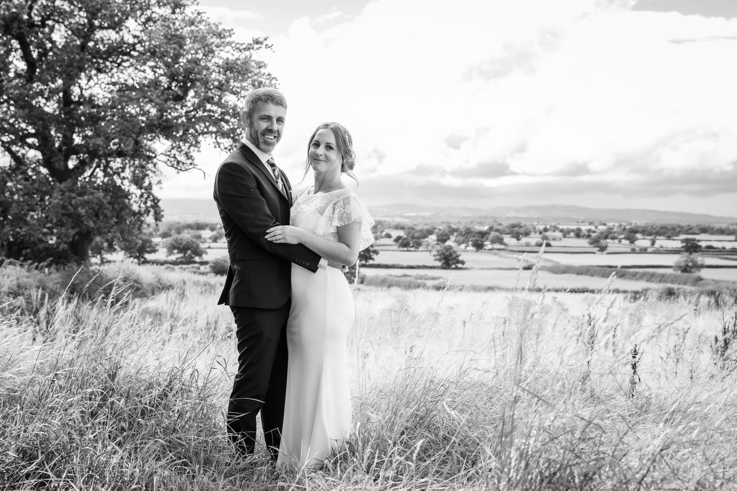 A wedding couple stands in a scenic field with rolling hills and countryside views in the background.