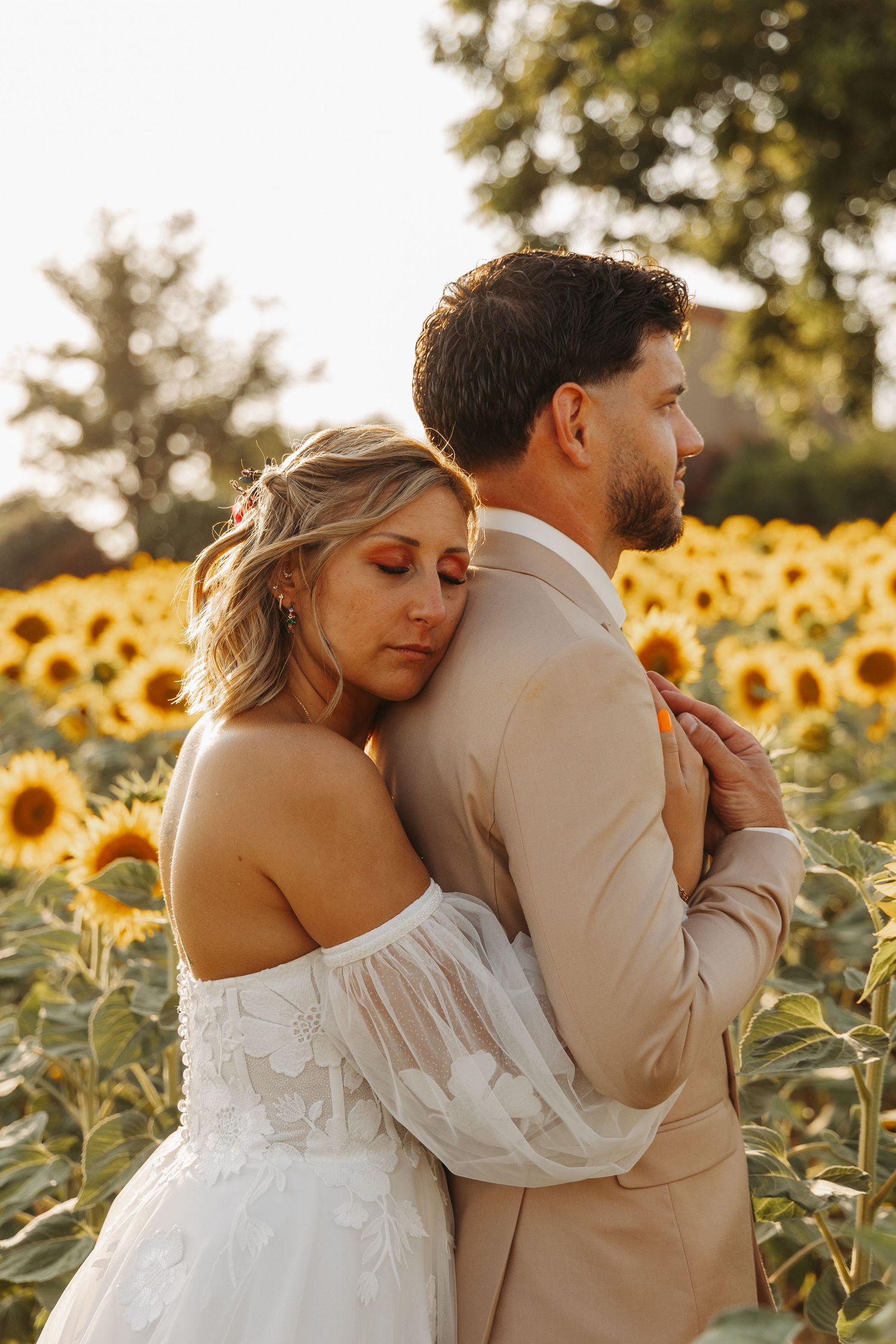 Bride resting her head on grooms back while embracing in a vibrant sunflower field at sunset.