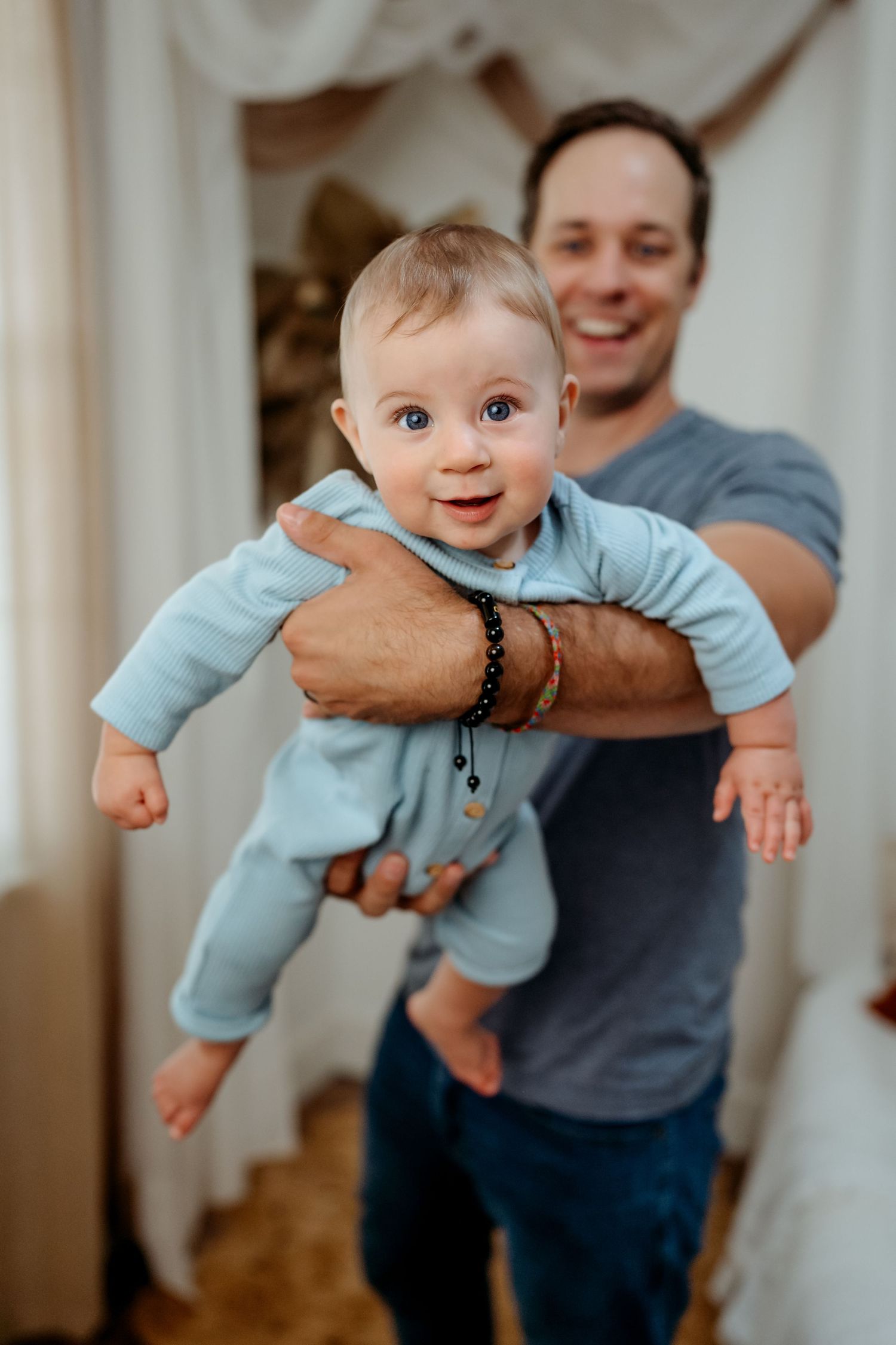 A baby wearing light blue pajamas smiles while being held up in the air at home.