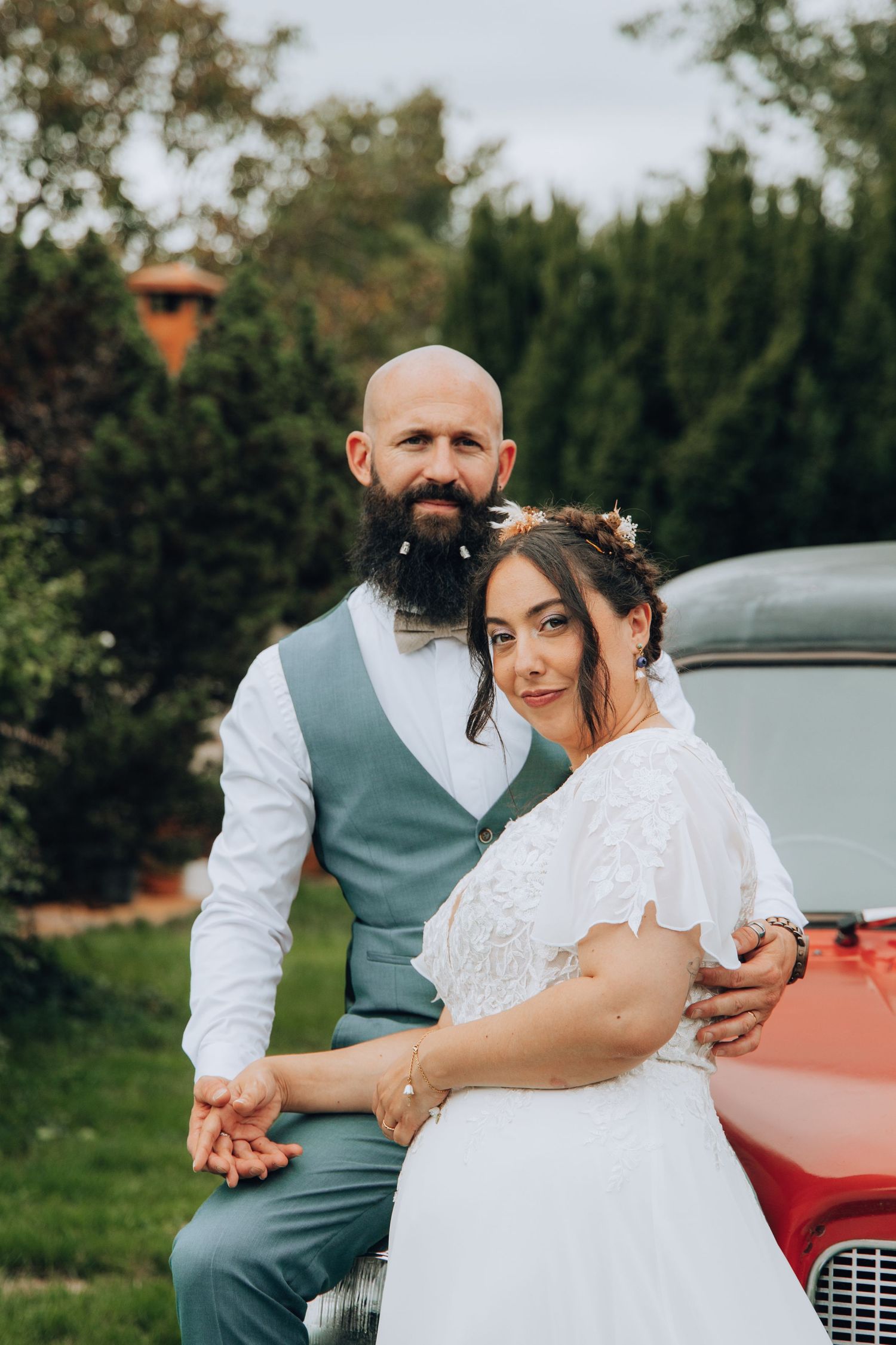 Bride and groom posing by a vintage red car, groom in gray vest with bow tie, bride in lace wedding dress.