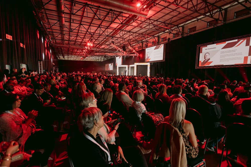 Large audience seated in a dimly lit conference hall watching a presentation on screen.