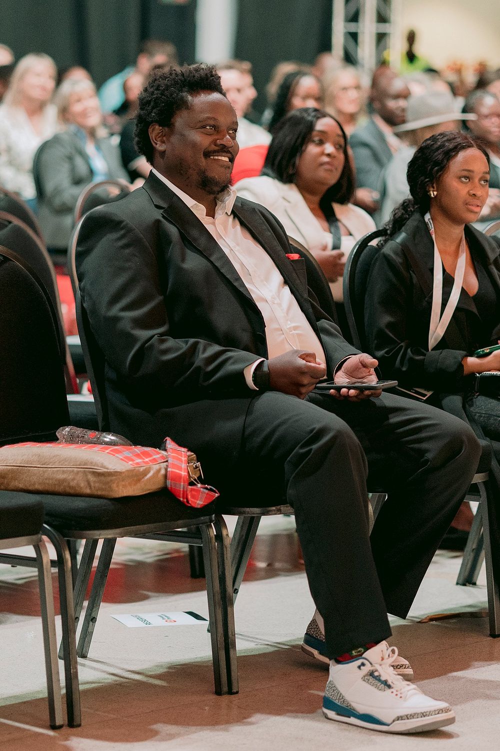 Smiling man in black suit and sneakers sits at a conference event with audience behind him.