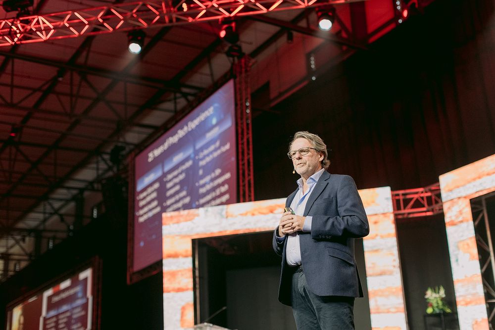Business speaker presenting on stage at a conference with large illuminated screens displaying data behind him.