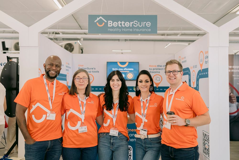 Five BetterSure team members in orange shirts smiling at their exhibition booth.