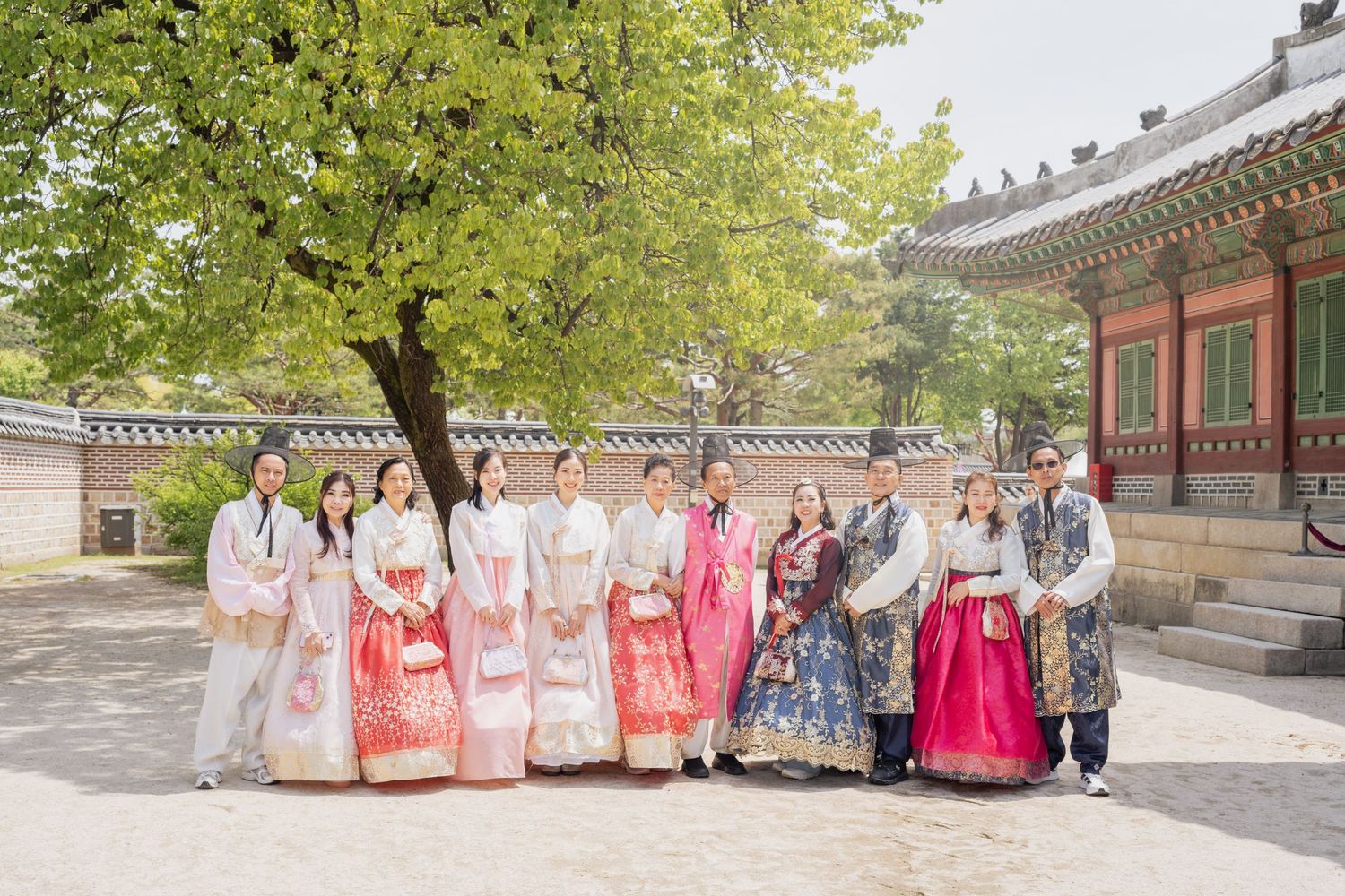 A group wearing colorful traditional Korean hanbok outfits stands together in a historic courtyard setting.