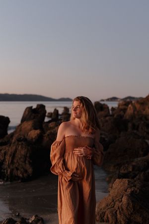 Figure in flowing peach dress poses gracefully among coastal rocks at sunset with ocean view in background.