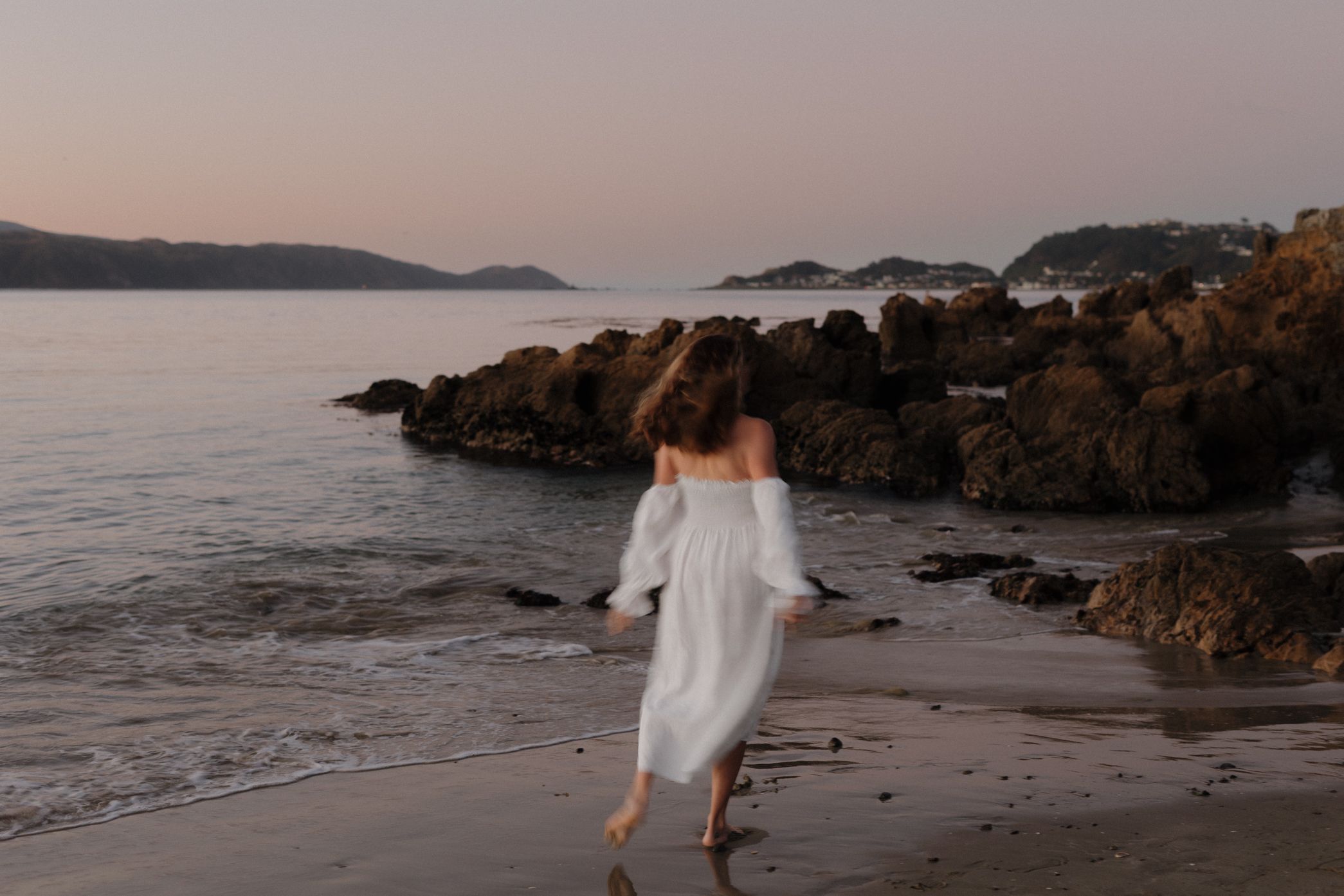 A person in a flowing white dress walks along a rocky beach at sunset with mountains in the distance.