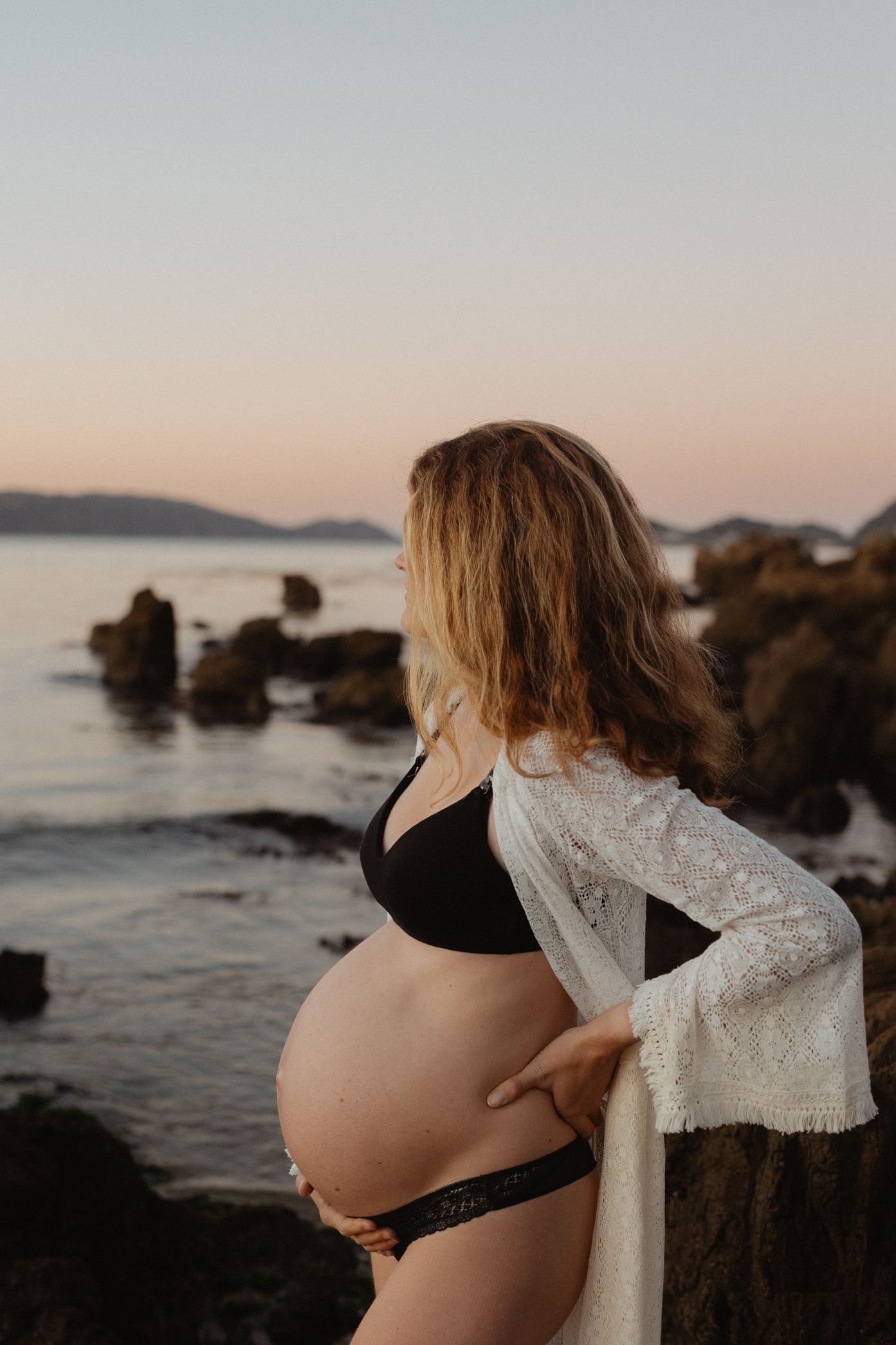 Pregnant woman in black swimwear and lace cover-up stands on rocky beach at sunset with ocean and mountains in background.