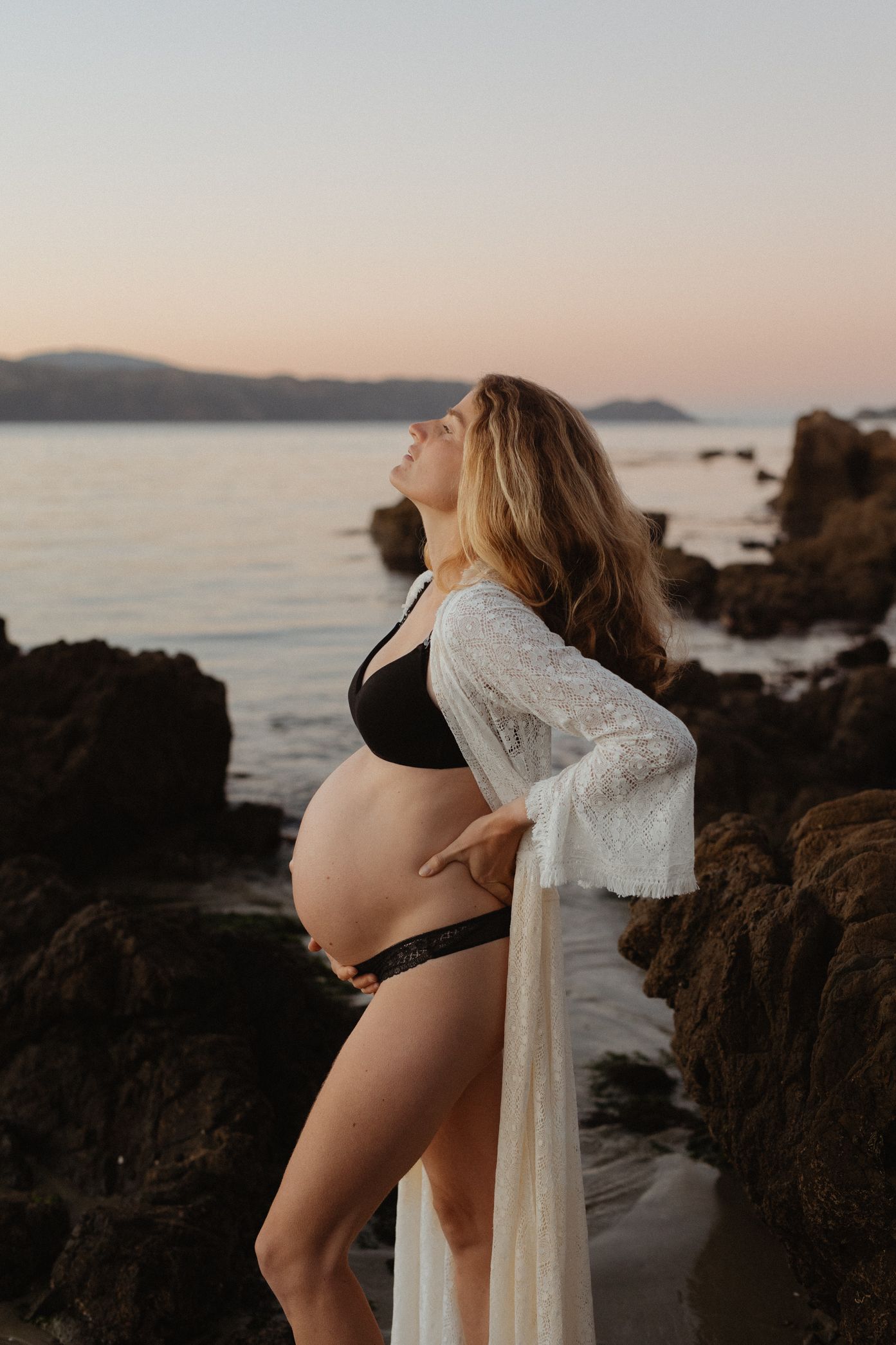 Pregnant figure in black swimwear and white lace robe poses on rocky beach at sunset with mountains in background.
