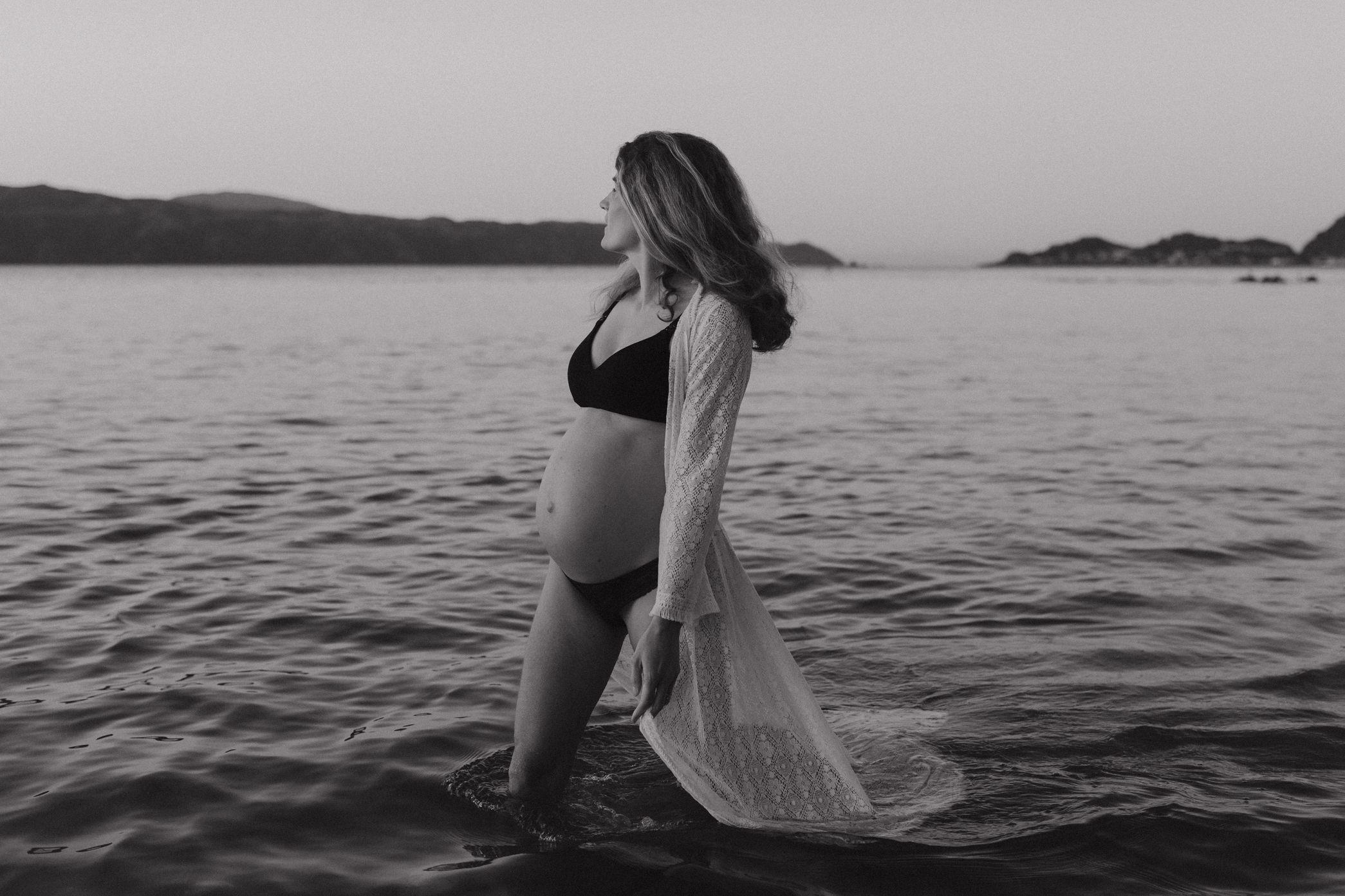 Black and white maternity photo of an expectant mother standing in water wearing a bikini and flowing sheer cover up.