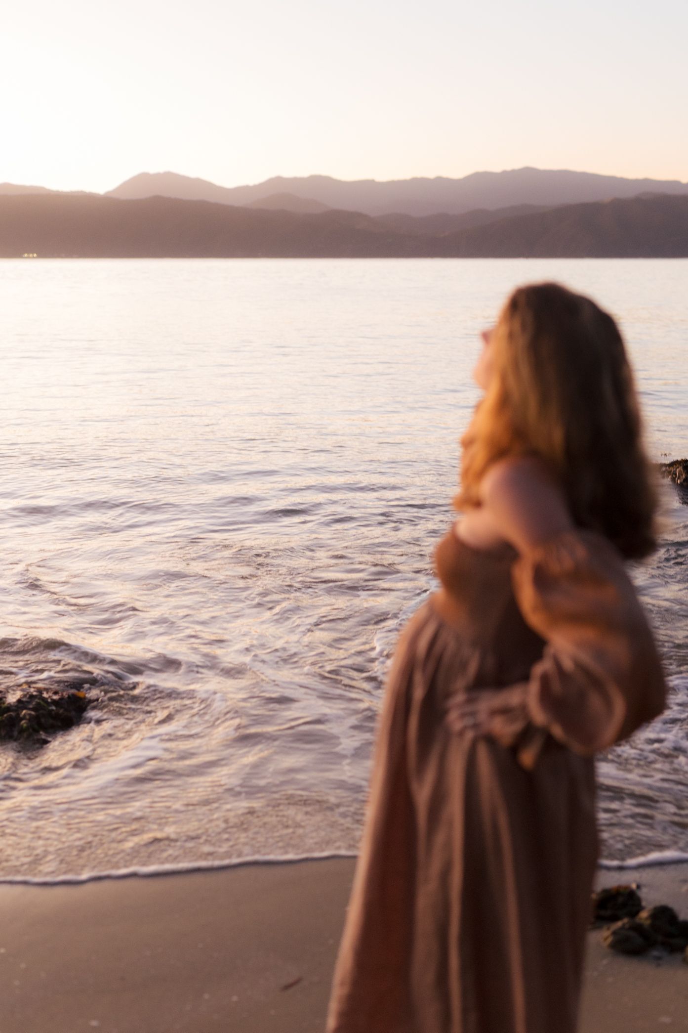 A person in a brown dress stands on a beach at sunset with mountains and ocean waves in the background.