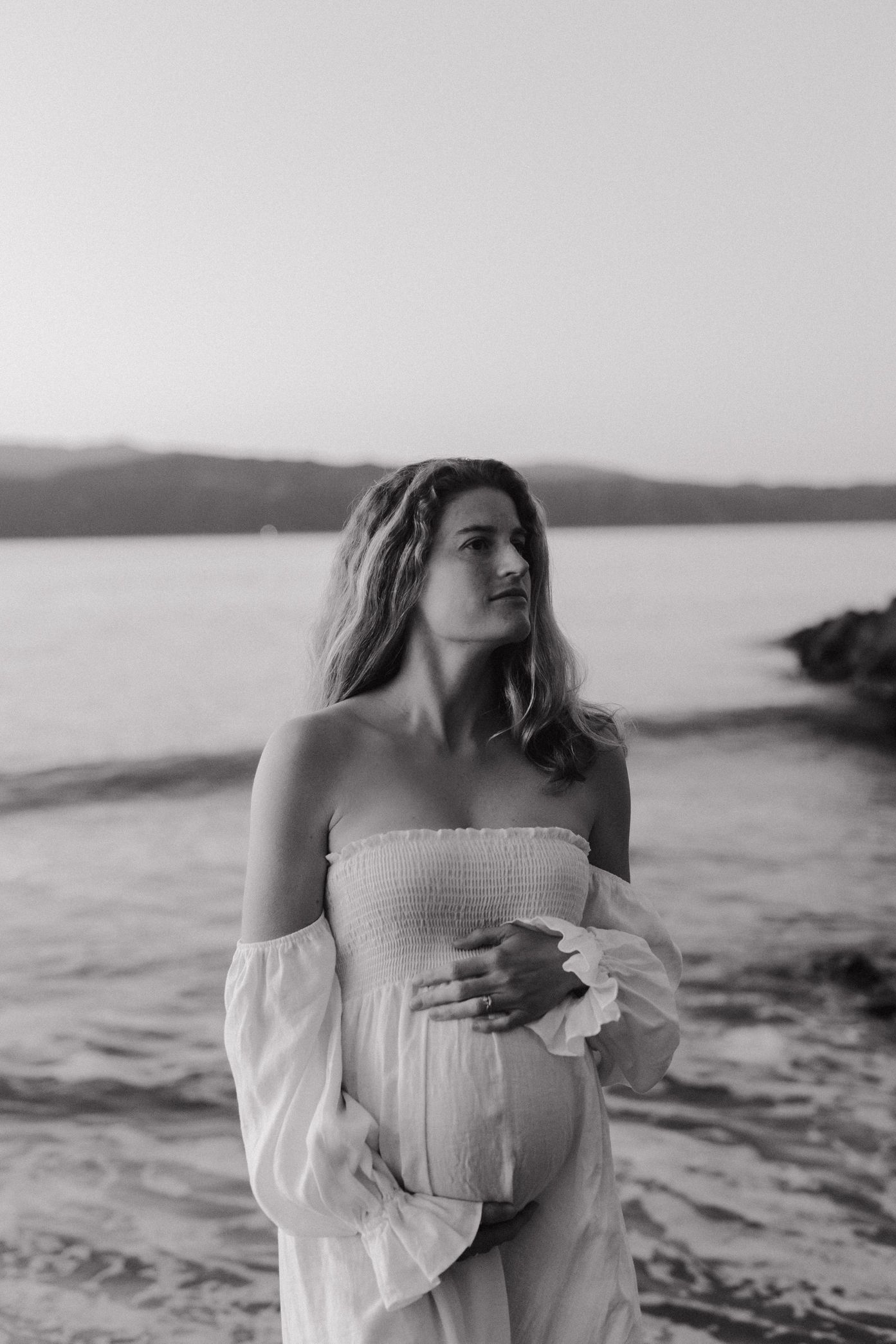 A person in a white off-shoulder dress poses thoughtfully by the ocean at sunset in black and white photography.