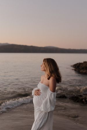 A person in a white flowy dress stands contemplatively by the ocean shore at sunset.