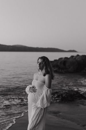 A person in a flowing white dress stands on a rocky beach at sunset with mountains in the distance.